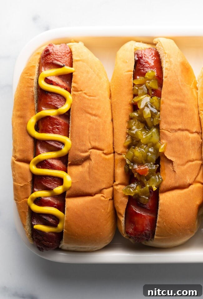 A top down view of two hot dogs on a white plate. One hot dog has zig zag mustard and the other one has relish.