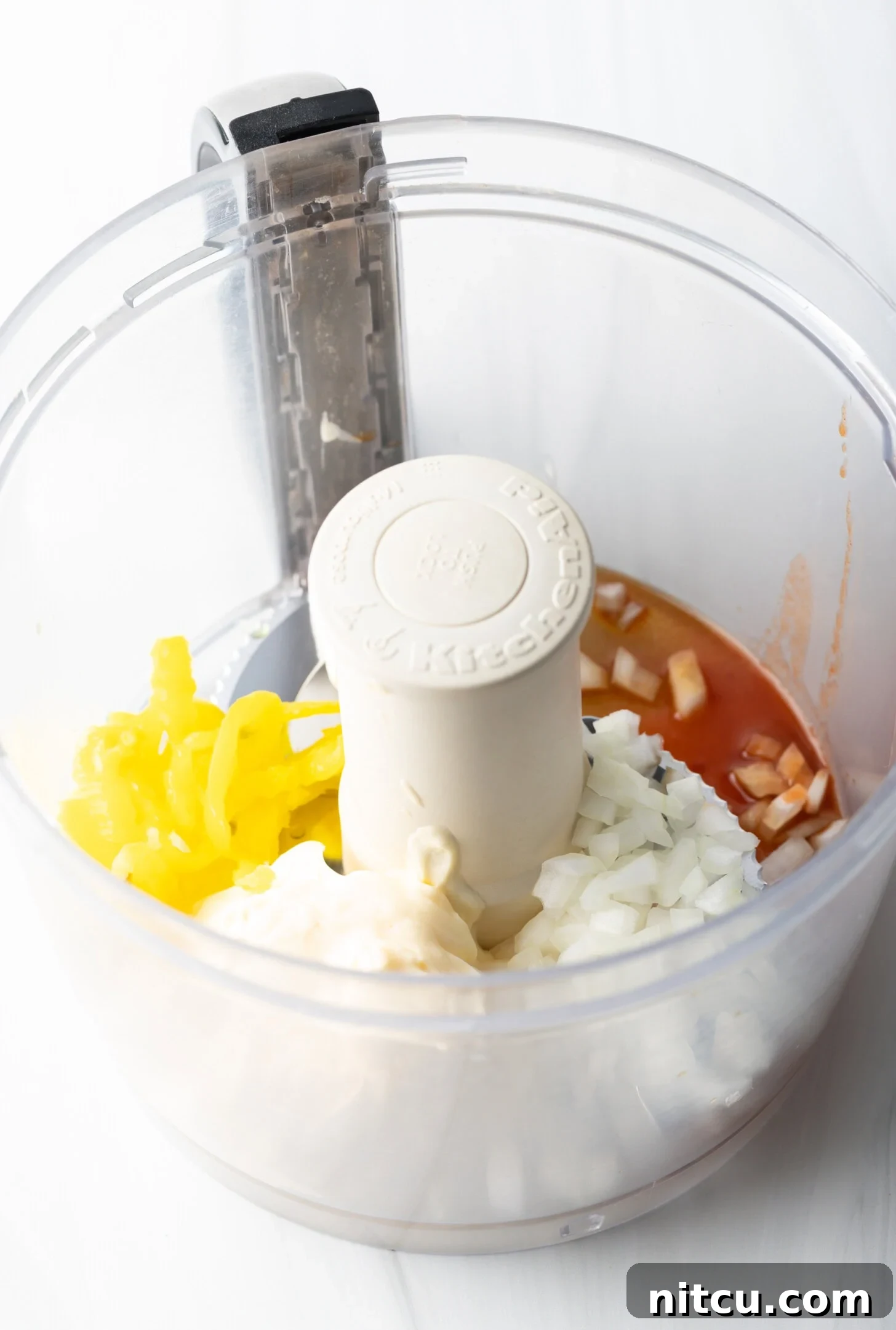 A vibrant close-up shot of chopped onions, bright yellow banana peppers, and a dash of fiery hot sauce, all contained within the clear bowl of a food processor, ready for blending.