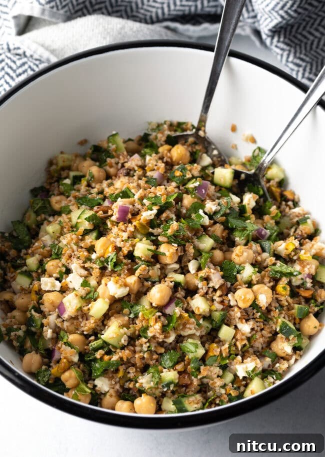 Large white bowl filled with bulgur wheat, veggies, and feta salad. Two metal salad spoons are in the bowl as well.