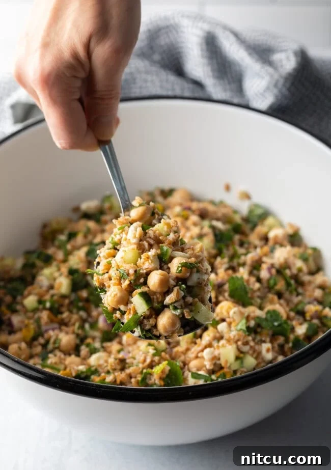 Hand with metal spoon scooping a serving of salad from a white bowl filled with bulgar wheat, veggies, and feta salad.