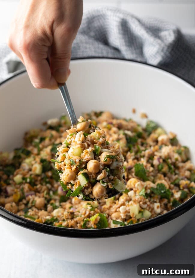 Hand with metal spoon scooping a serving of salad from a white bowl filled with bulgur wheat, veggies, and feta salad.