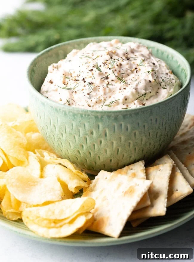 A vibrant green bowl filled with creamy clam dip, beautifully arranged on a white plate and surrounded by an assortment of crispy potato chips and crackers.