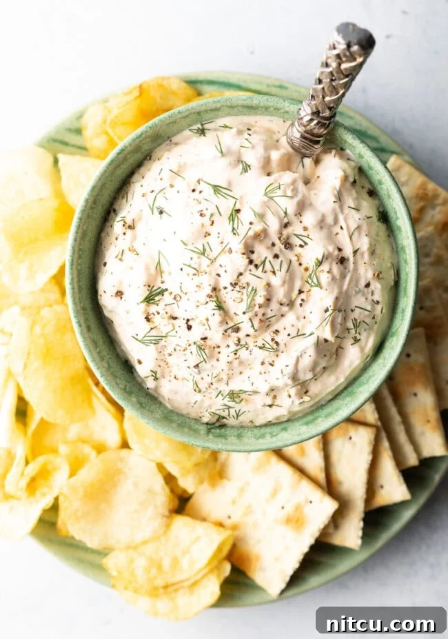 A top-down view captures a green bowl overflowing with creamy clam dip, carefully placed on a white plate. It is encircled by an inviting spread of various potato chips and crisp crackers, ready for dipping.