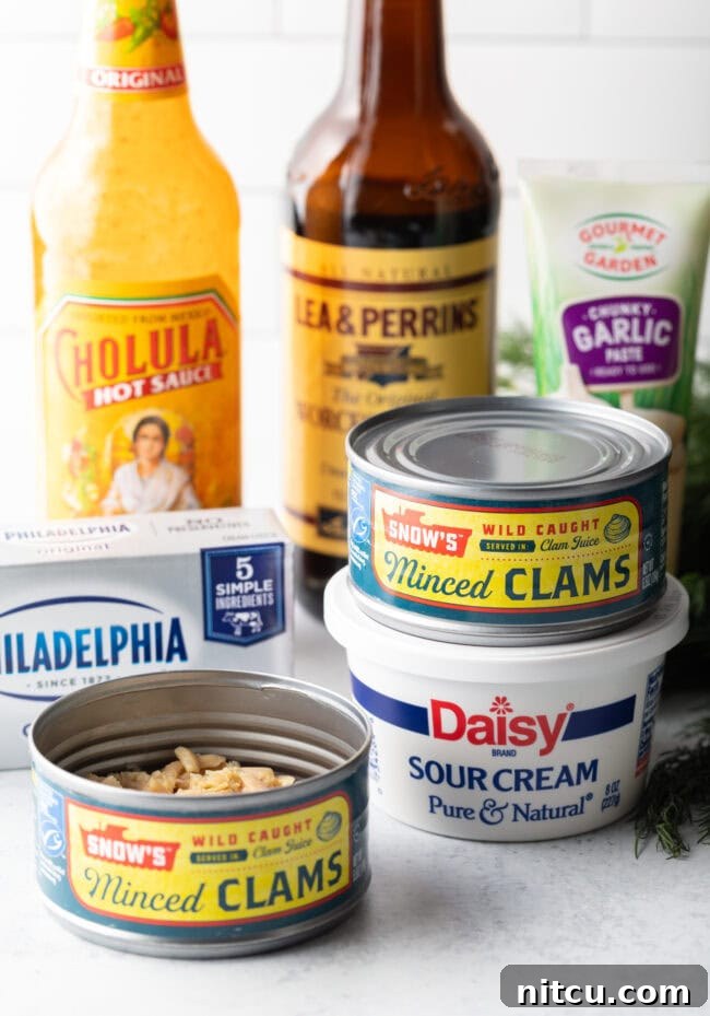 A perfectly arranged shot showcasing all the essential recipe ingredients. Featured are a bottle of Cholula hot sauce, a bottle of Worcestershire sauce, two cans of minced clams, a block of cream cheese, and a container of sour cream, all waiting to be transformed.