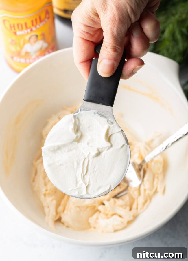 A hand holds a measuring cup, pouring creamy sour cream into a white bowl already containing a seasoned cream cheese mixture, signifying the next step in the recipe preparation.