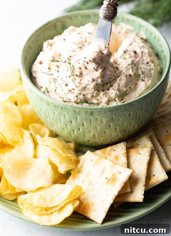 A beautifully presented green bowl brimming with creamy clam dip, sitting on a pristine white plate, encircled by a generous selection of potato chips and crackers.