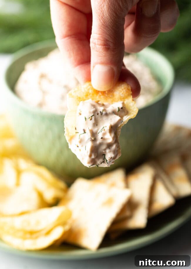 A hand skillfully scoops a generous portion of homemade clam dip onto a crisp potato chip, holding it up towards the camera in an inviting gesture.