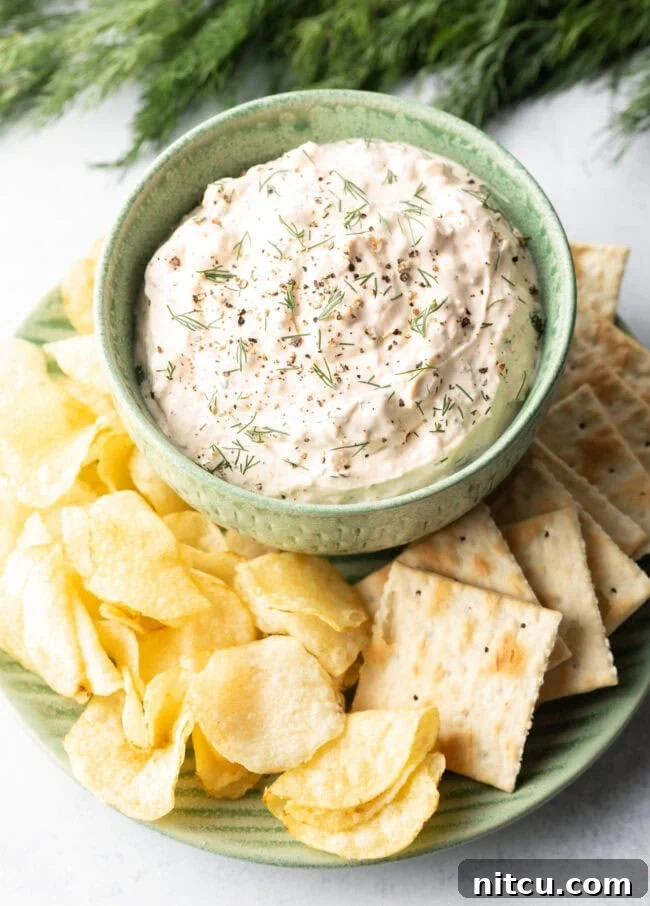 A top-down perspective highlights a inviting green bowl, generously filled with the best clam dip recipe, artfully placed on a white plate and encircled by a variety of crispy potato chips and crackers.