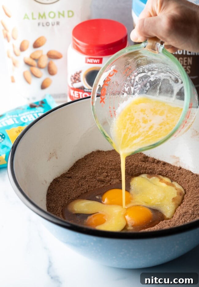 A hand pouring melted butter from a glass measuring cup into a bowl containing the dry ingredients and eggs, beginning the mixing process.