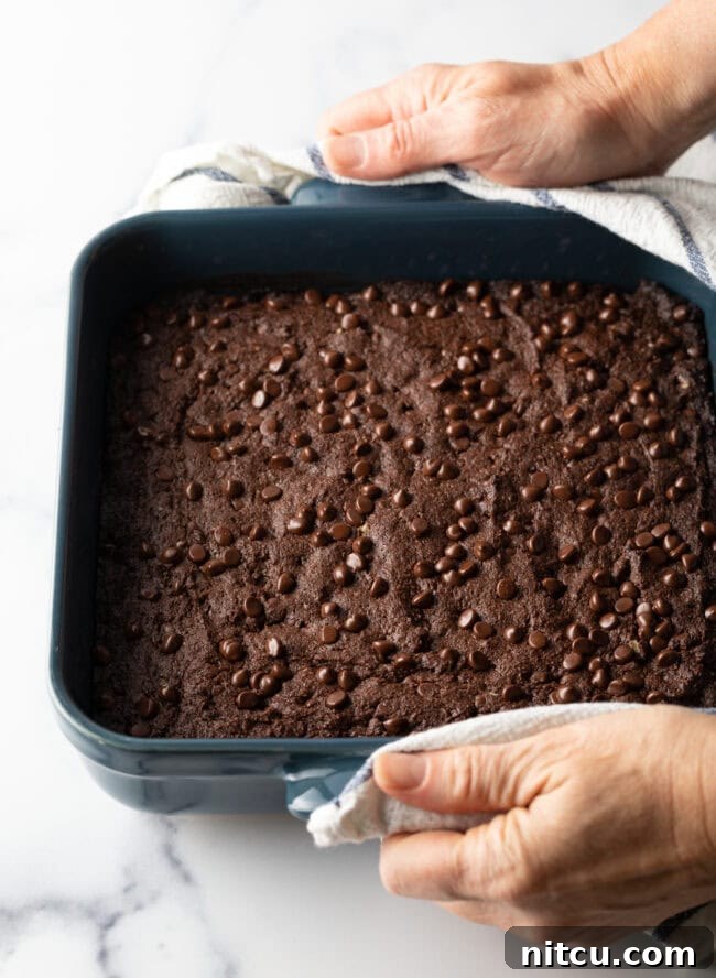 A hand holding a square pan of freshly baked brownies, dotted with melted chocolate chips, just out of the oven.