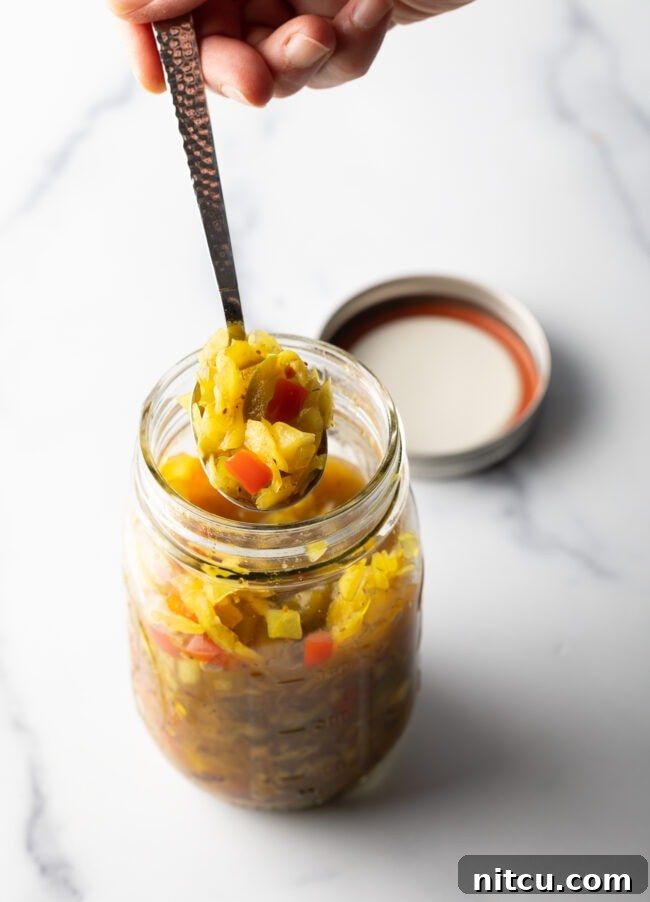 Top down view glass jar of chow chow. The lid is off and to the right side, a metal spoon is in the jar showing a scoop to camera.