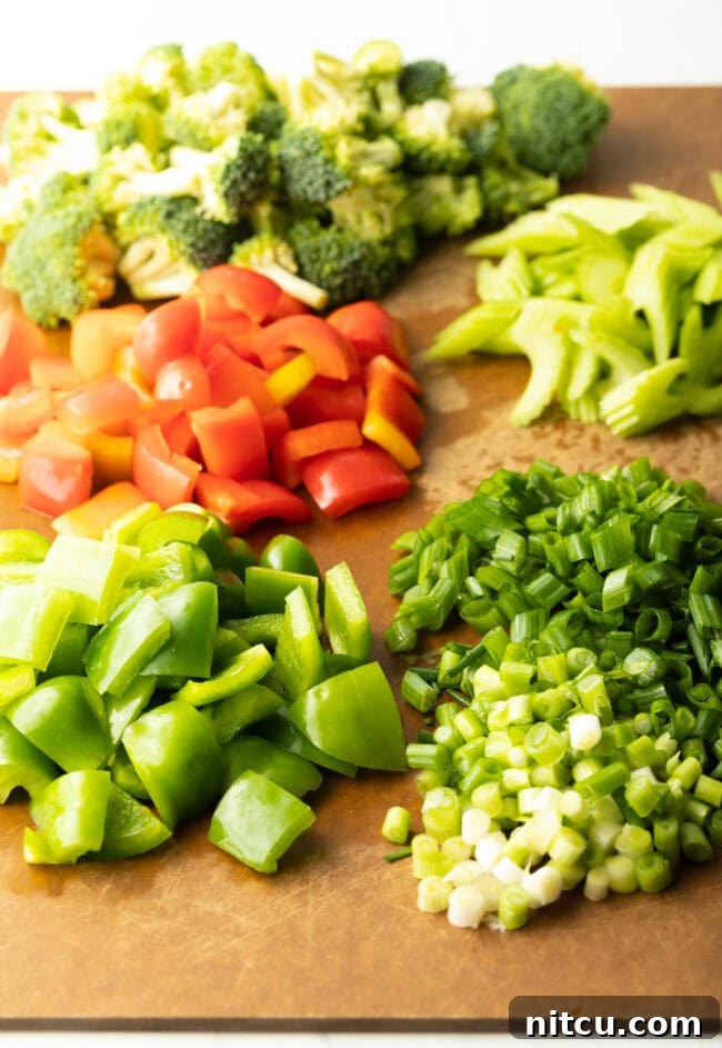 A cutting board laden with neatly organized piles of chopped green bell pepper, red bell pepper, scallions, celery, and broccoli florets, showcasing precise preparation.