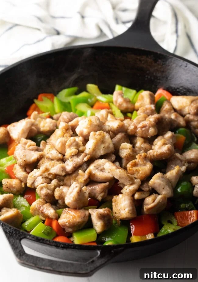Cooked chicken pieces are being added back into the skillet with sautéed vegetables, ready to be coated in the sauce.