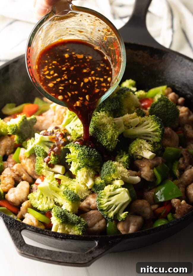 The savory Hunan sauce is being poured over the colorful medley of chicken and vegetables in a sizzling skillet, creating a rich glaze.