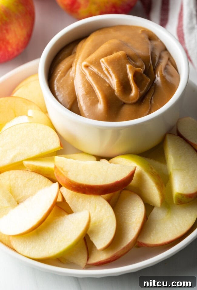 A pristine white bowl brimming with creamy caramel apple dip, elegantly presented on a white plate, encircled by a vibrant array of freshly sliced apples.