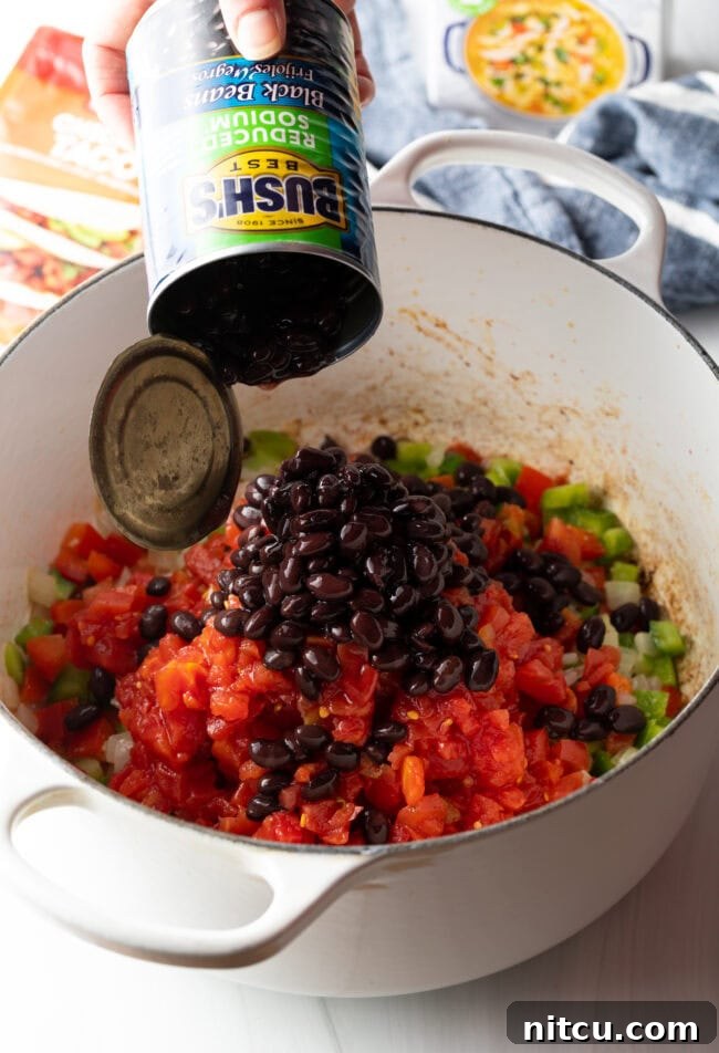 Hand adding canned black beans to pot with chopped veggies.