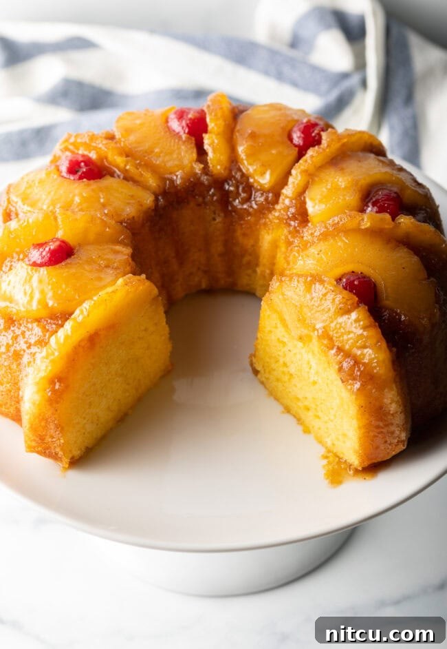 A pineapple upside down Bundt cake on a white cake stand with a significant slice removed, offering a clear view of the fluffy yellow cake inside.