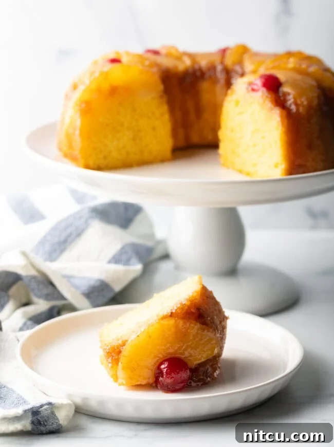 A baked pineapple upside down Bundt cake displayed on a white cake stand with a slice removed, revealing the soft yellow interior. A separate slice rests on a white plate in the foreground.