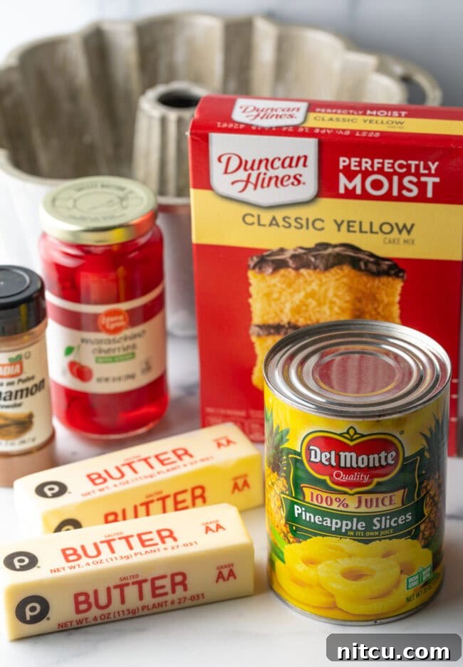 Various recipe ingredients including a box of yellow cake mix, a jar of maraschino cherries, a can of pineapple, two sticks of butter, and ground cinnamon spice.