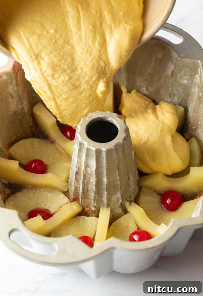 Pouring the smooth yellow cake batter over the meticulously placed pineapple slices and cherries at the bottom of the Bundt pan.