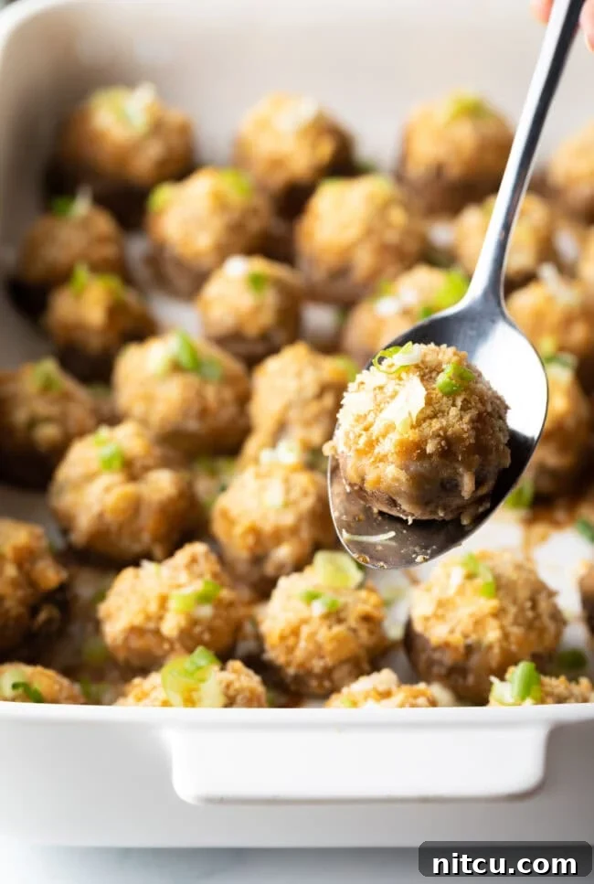 Two dozen seafood stuffed mushrooms in a white baking dish. A metal spoon is showing 1 mushroom to camera.