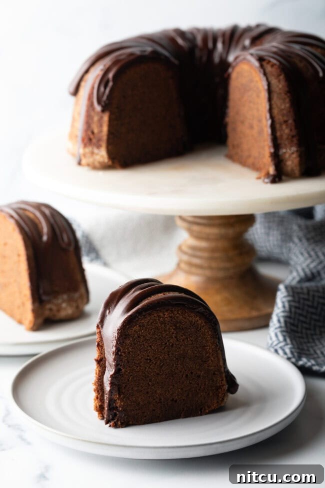 Chocolate Glaze for Pound Cake - Thick slice of chocolate sour cream pound cake with drizzle of glaze on a white plate. The full cake is on a white cake stand in the background.
