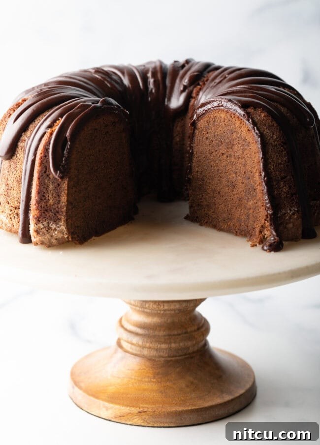 Chocolate sour cream bundt cake with a thick chocolate glaze drizzled on top. The cake is on a white stand and has a portion removed to show the tender inside crumb.