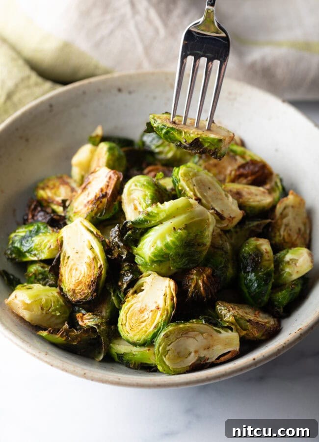A fork holding a single air-fried Brussels sprout half over a white bowl of additional crispy sprouts.