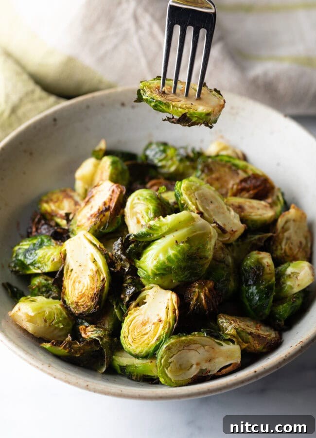 A fork lifts a perfectly cooked, halved air fryer Brussels sprout from a white bowl, showing its crispy edges.