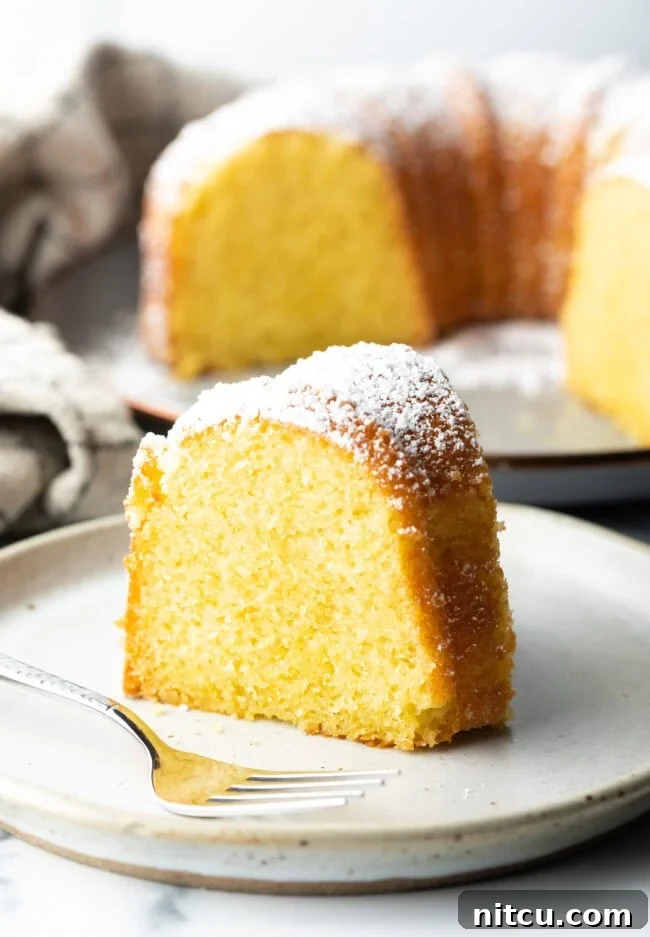 A perfectly baked slice of vibrant yellow lemon olive oil cake, glistening with moisture, presented on a pristine white plate, inviting a taste of its rich citrus aroma and tender crumb.