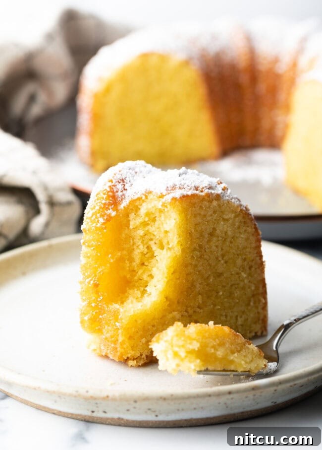 A close-up shot of a single slice of moist yellow cake on a white plate, with a fork gently cutting into a bite, highlighting its soft texture and inviting crumb.