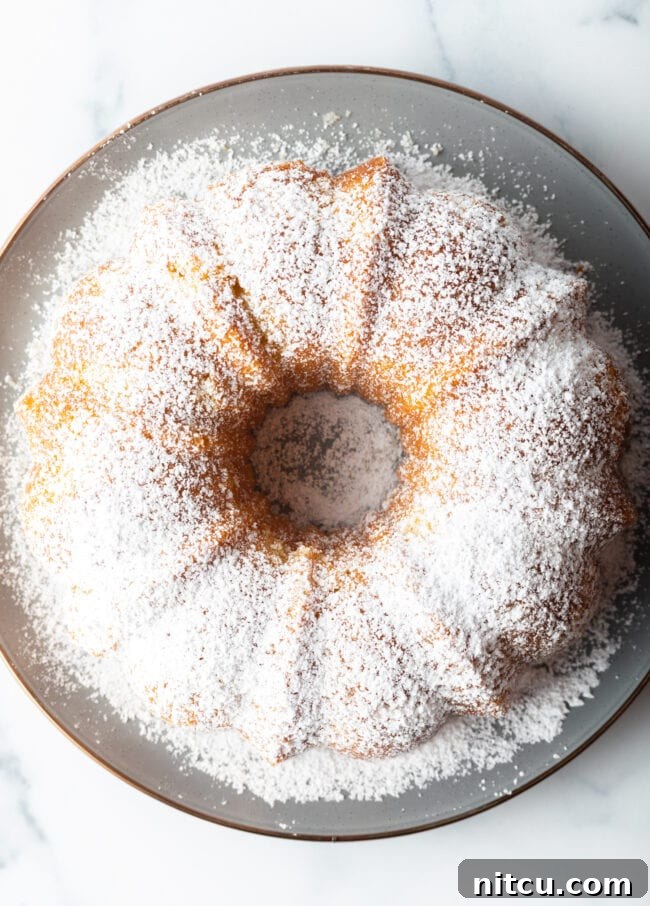 A stunning bundt cake, golden-brown and elegantly dusted with powdered sugar, awaits slicing on a white plate, showcasing the beautiful form achievable with this recipe.