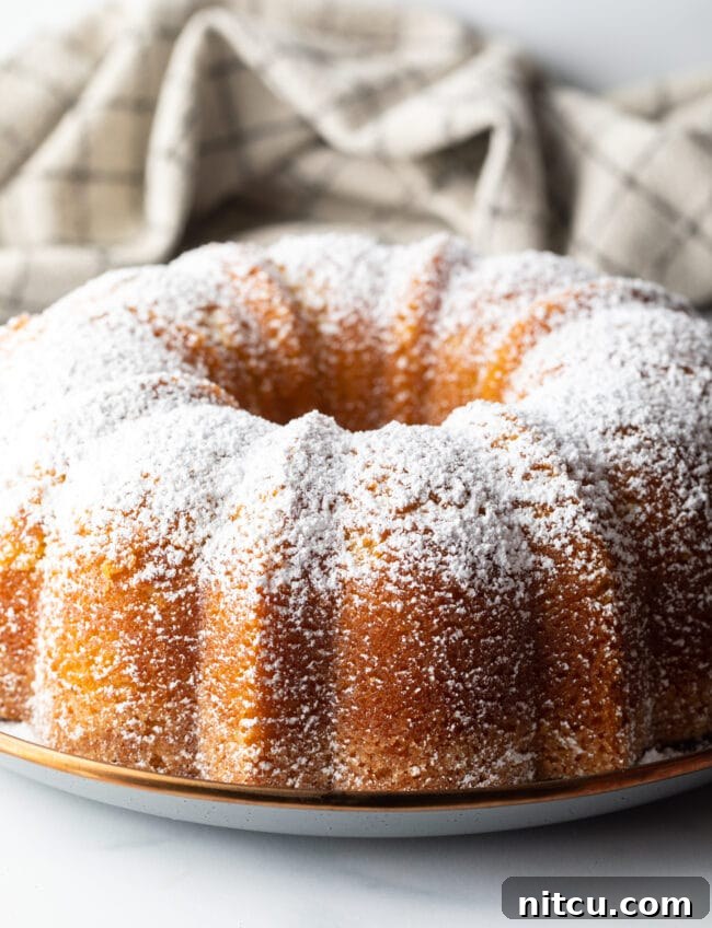 A freshly baked lemon olive oil bundt cake, elegantly dusted with fine powdered sugar, rests on a white plate, ready to be admired and enjoyed.