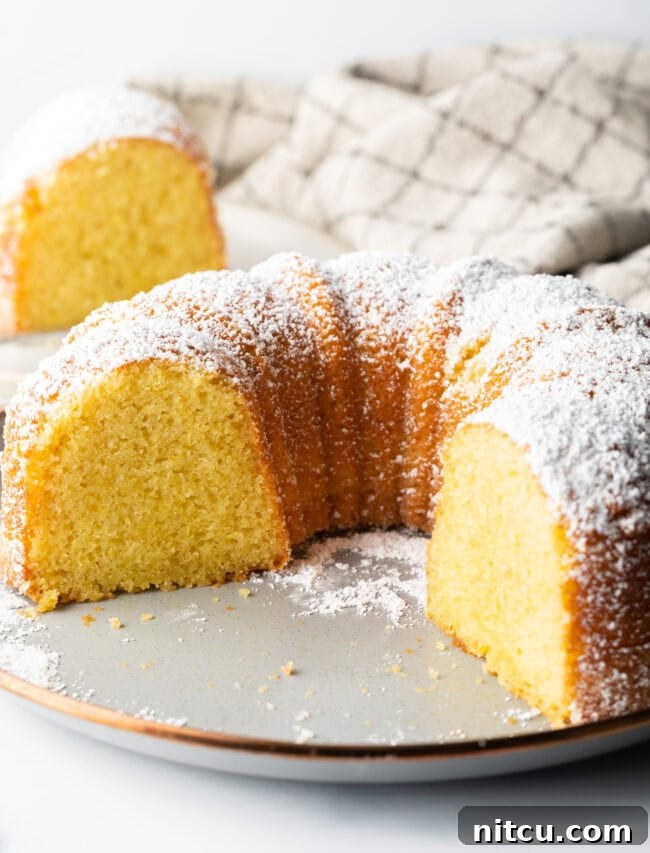 A generous slice of lemon olive oil bundt cake has been removed from the full cake, revealing the incredibly moist, tender, and bright yellow interior crumb.