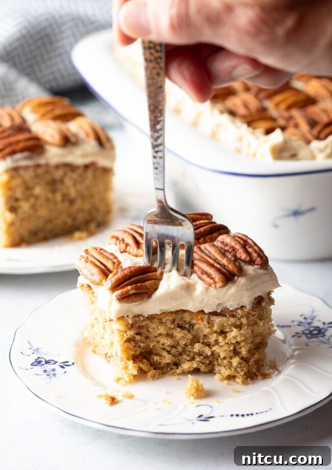 Square of brown butter cake on a plate with pecan halves on top of the white frosted cake. A fork is taking a bite of cake.