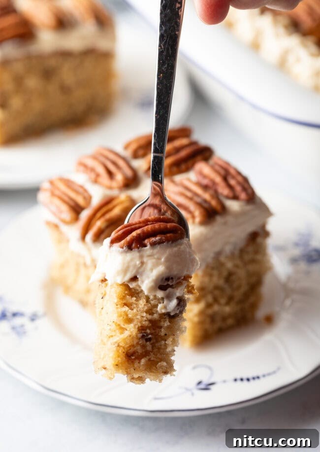Square of brown butter cake on a plate with pecan halves on top of the white frosted cake. A fork is taking a bite of cake.