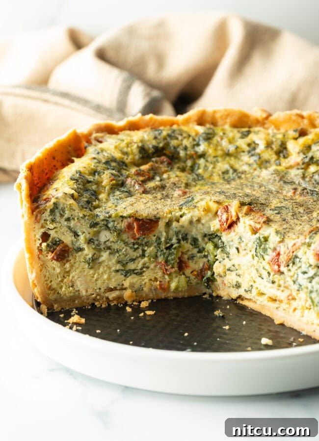 A beautifully baked deep dish veggie quiche on a white plate, with a large section removed to reveal the rich, vegetable-filled interior and perfectly cooked custard.