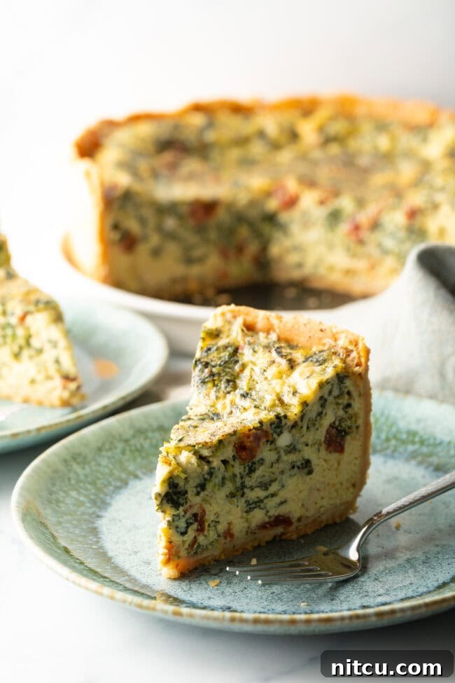 A close-up of a tall, perfect slice of quiche on a light blue plate, with a fork beside it, highlighting its flaky crust and rich, varied filling.