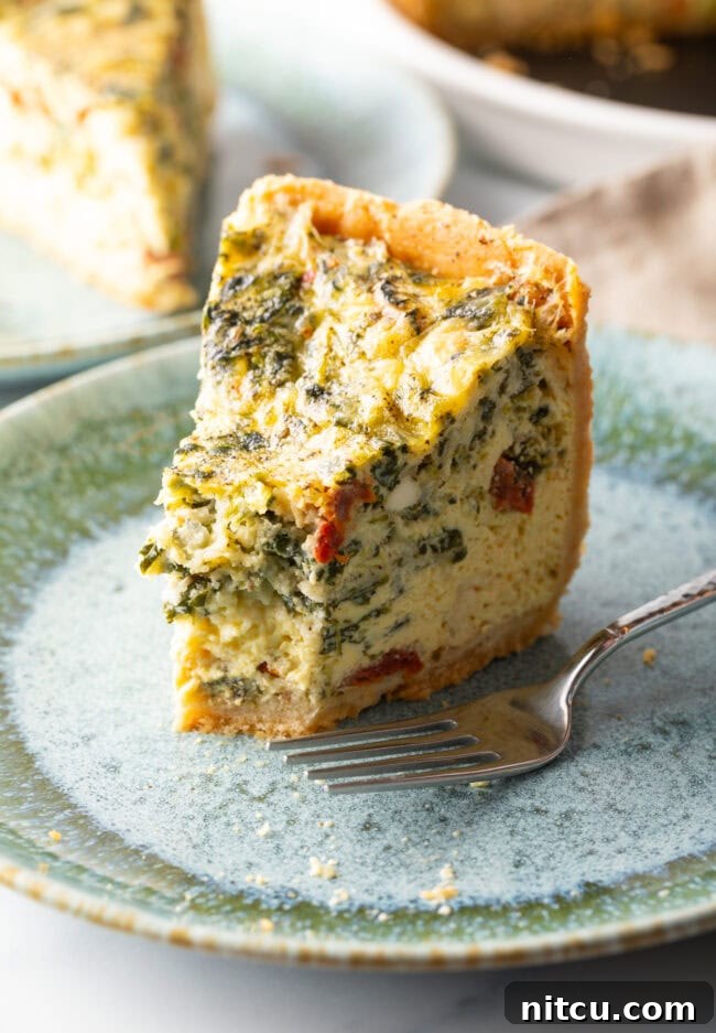 A tall, perfectly baked slice of quiche on a light blue plate, with a small bite taken from its tip and a fork resting nearby.