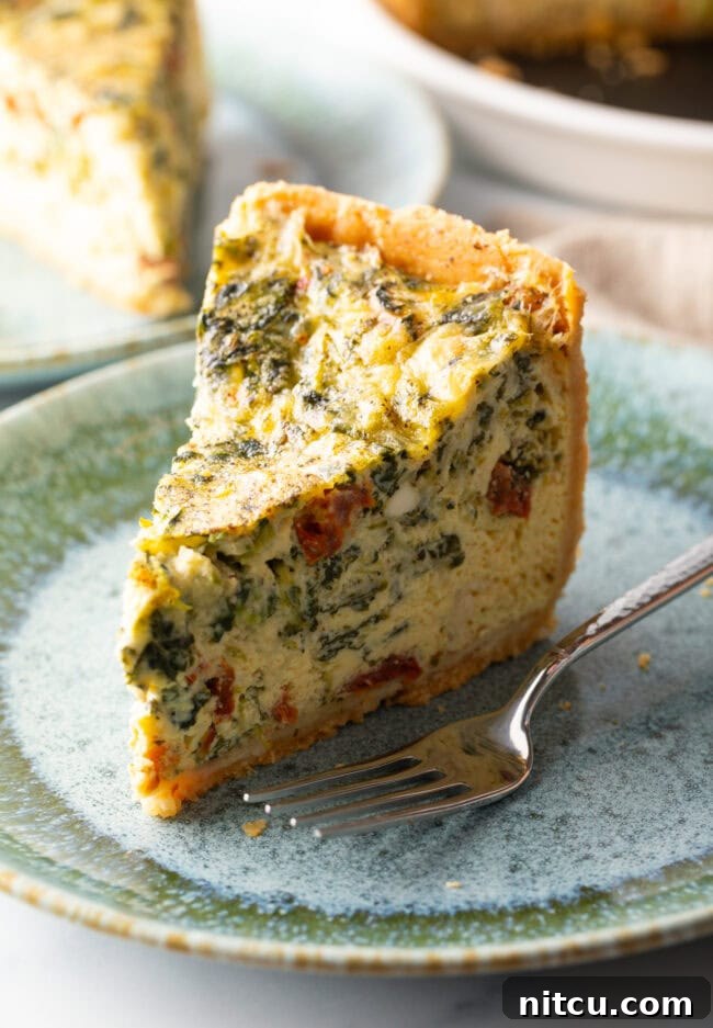 A tall, perfectly baked slice of vegetarian quiche rests on a light blue plate, with a fork positioned invitingly beside it. The visible layers showcase the flaky crust and the abundant vegetable filling.