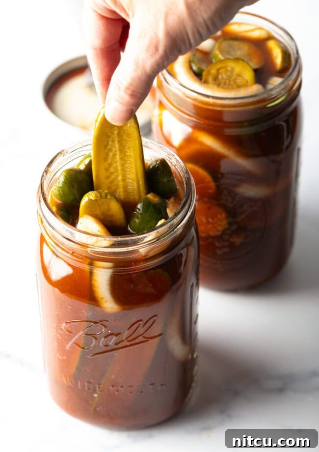 Two large mason jars of chamoy pickles. The tops are off of the jars, and a long pickle is being pulled out of one jar.