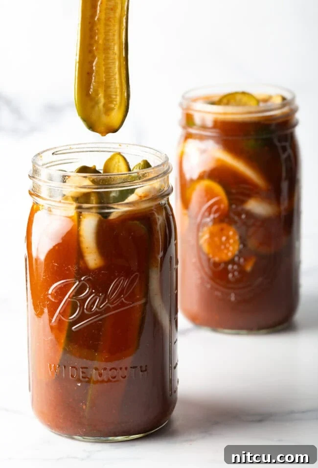 Two large mason jars of chamoy pickles. The tops are off of the jars, and a long pickle is being pulled out of one jar.