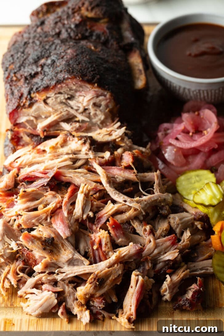Shredded smoked pork butt piled on a rustic cutting board, accompanied by a ramekin of homemade BBQ sauce, a stack of crispy dill pickles, and vibrant pickled red onions, ready to be served.