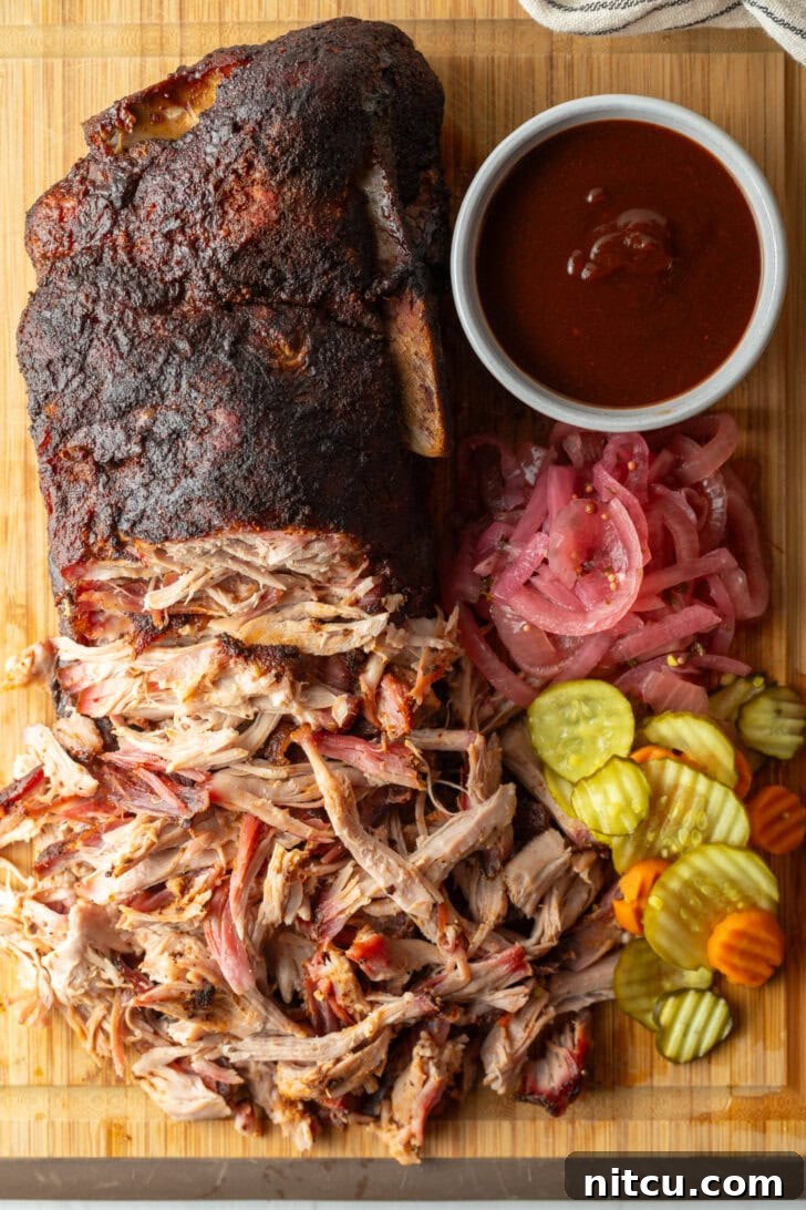 Close-up of shredded smoked pork butt on a wooden cutting board, garnished with a side of savory BBQ sauce, crunchy pickles, and tangy pickled red onions.