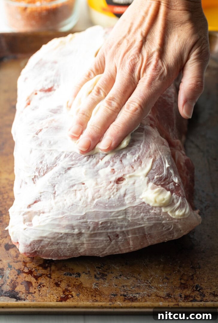 The raw pork shoulder, glistening with a thin coat of mayonnaise, preparing for the dry rub application.