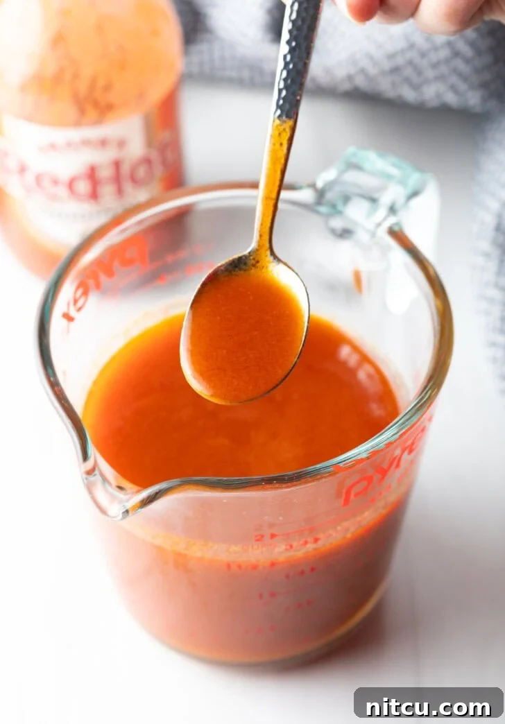 Spoon drizzling buffalo sauce from a large glass measuring cup.