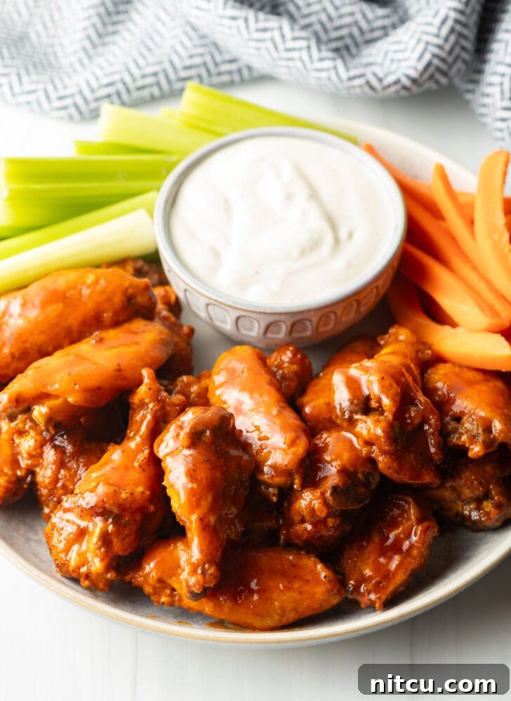 Plate of saucy buffalo chicken wings. On the plate is also a ramekin of ranch dressing, a pile of carrot sticks, and a pile of celery sticks.