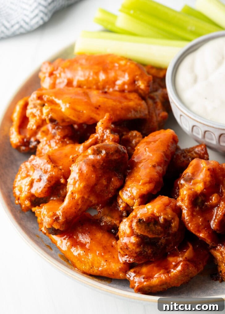 Plate of saucy buffalo chicken wings. On the plate is also a ramekin of ranch dressing, a pile of carrot sticks, and a pile of celery sticks.