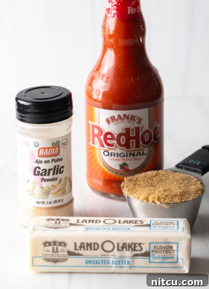 Recipe ingredients on a white backdrop: Bottle of Frank's RedHot, metal scoop of brown sugar, container of garlic seasoning, and a stick of butter.