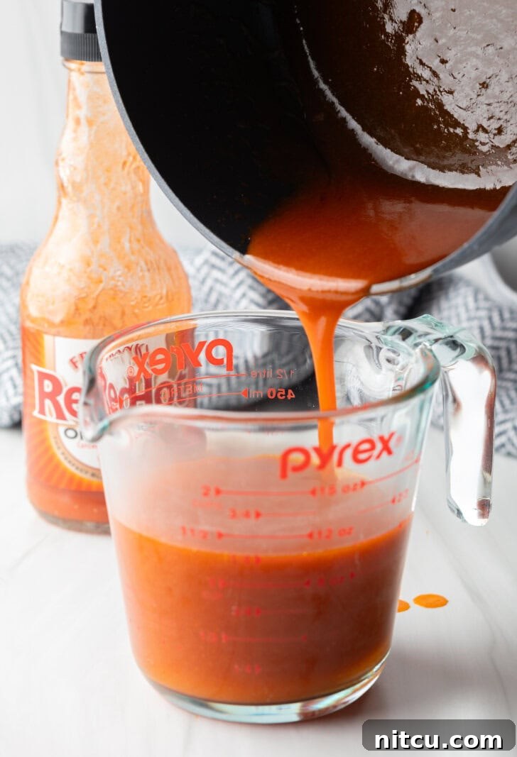 Pouring homemade buffalo sauce from a black sauce pot into a large glass measuring cup.